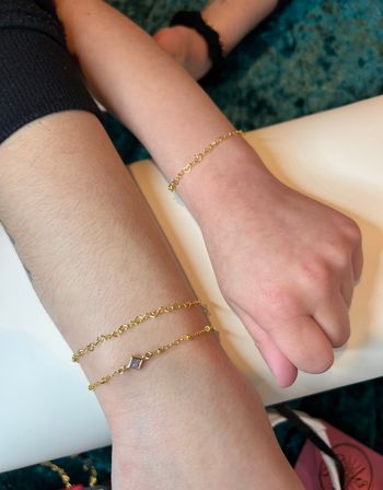 Close-up of two wrists resting on a white surface wearing delicate gold chain bracelets, one wrist layered with a small square charm and the other with a single heart-link chain, teal carpet background