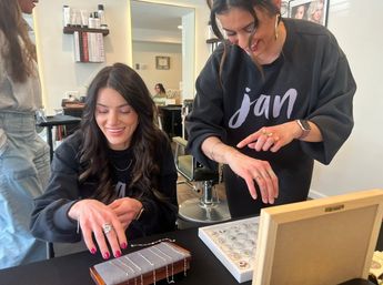 Smiling woman trying on rings during a jewelry-styling moment at a beauty salon, stylist pointing to bracelets and rings on a display tray with mirror and salon chairs in the background.