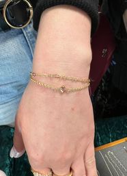 Close-up of a hand at a jewelry counter wearing two delicate layered gold bracelets—one twisted chain and one with a small square crystal charm, with denim and a black belt visible in the background.