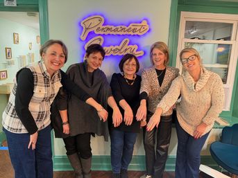 Five smiling women in a boutique jewelry studio proudly showing matching chain bracelets on their wrists beneath a glowing "Permanent Jewelry" neon sign, casual indoor group photo.