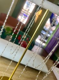 Gleaming silver-tone chain necklaces hanging from a gold T-bar jewelry display, boutique interior with colorful floral seating and sunlit window blurred in the background
