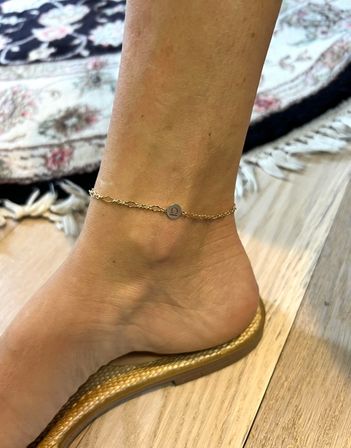 Close-up of an ankle wearing a delicate gold chain anklet with a small round charm, foot in a woven gold sandal on a light wood floor beside a patterned area rug.