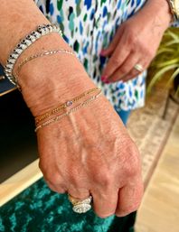 Close-up of a hand and wrist wearing stacked gold and silver chain bracelets with a tiny purple gemstone and a sparkling diamond cluster ring, set against a blue-green patterned blouse and pink nail polish.