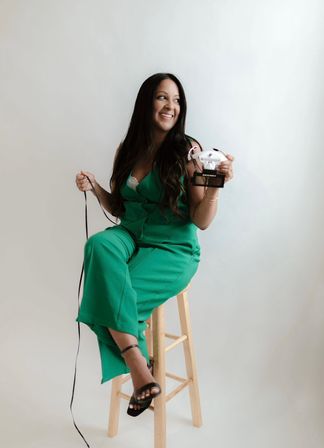Smiling woman in a green jumpsuit seated on a wooden stool in a studio, holding a white VR headset and cable against a neutral background