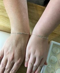 Two wrists, adult and child, wearing matching delicate silver chain bracelets with small bar and heart links, resting on a light wood table beside a jewelry tray.
