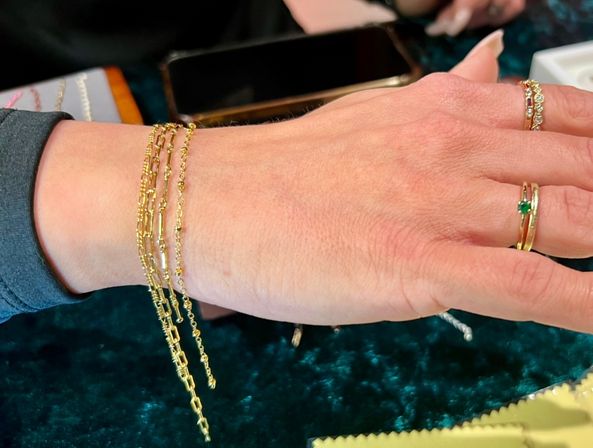 Close-up of a hand wearing delicate gold chain bracelets and stacked gold rings with an emerald and small diamonds, resting on dark teal velvet jewelry display with a smartphone nearby