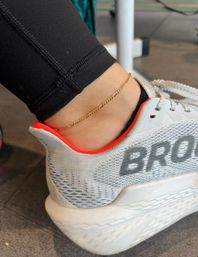 Close-up of ankle wearing a delicate gold chain anklet over black leggings next to a white and gray running shoe with red trim on a gym floor.
