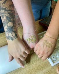 Three hands on a wooden table showing gold temporary tattoos reading "Bride" and "Bridal Tribe", delicate chain bracelets and a bold floral forearm tattoo, with a jewelry display tray nearby — bridal party bachelorette vibe.