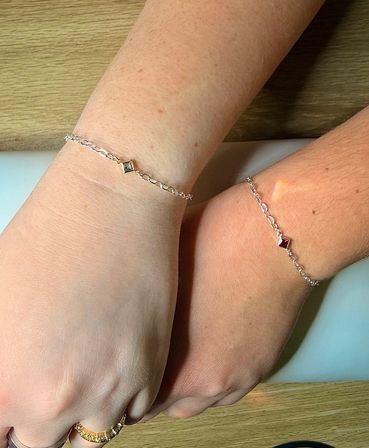 Close-up of two wrists resting on a light wood surface wearing paired minimalist silver chain bracelets with small square gemstone charms