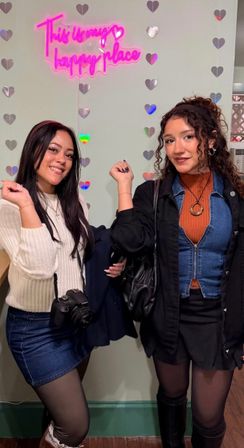 Two friends posing at a neon photo wall with a bright pink sign reading "This is my happy place" and hanging holographic hearts; one holds a DSLR camera, both wear denim and skirts in a cozy indoor selfie spot.