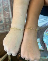 Close-up of two wrists side-by-side wearing matching delicate gold chain bracelets with open-heart links and small gemstone charms in a casual indoor/home setting