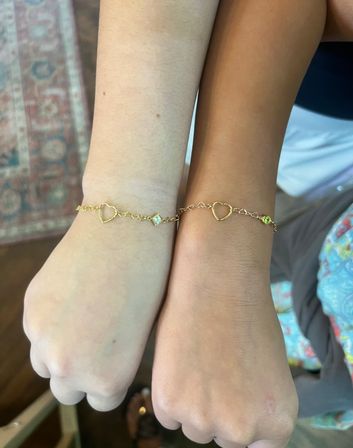 Close-up of two wrists side-by-side wearing matching delicate gold chain bracelets with open-heart links and small gemstone charms in a casual indoor/home setting