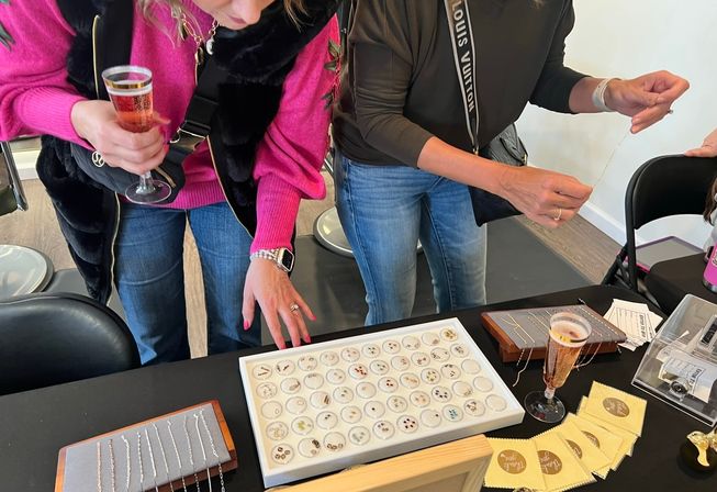 Two shoppers leaning over a jewelry display at a pop-up market, examining trays of delicate stud earrings and chain necklaces on a table with two sparkling wine flutes nearby.