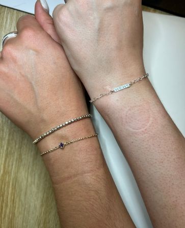 Close-up of two wrists side-by-side on a light wood surface wearing minimalist wrist jewelry: stacked gold and silver bracelets with a small purple gemstone and a delicate silver chain with a rectangular nameplate.