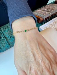 Close-up of a hand wearing a delicate gold chain bracelet with a small round green gemstone, photographed over teal velvet beside a tray of jewelry.