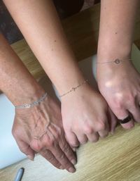 Three wrists resting on a light wood surface showcasing delicate bracelets: left silver chain with turquoise beads and peace charm, center thin gold chain with a small square gemstone, right silver chain with round disc charm and a black ring on the finger.