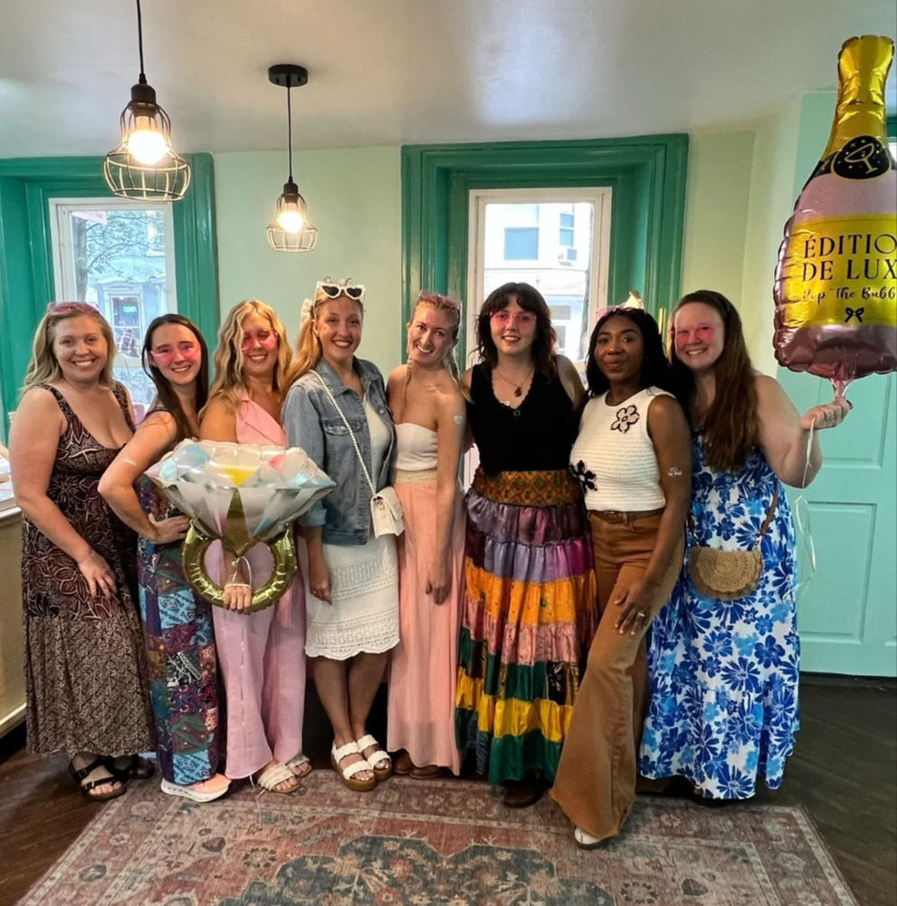 Group of eight women posing indoors in a teal-accented room for a celebratory gathering — colorful boho outfits, oversized diamond ring and champagne balloons, hanging pendant lights and patterned rug