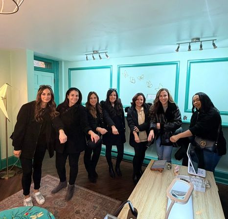 Seven women posing and extending their hands to show manicures inside a teal-accented nail studio with a reception desk and lamp