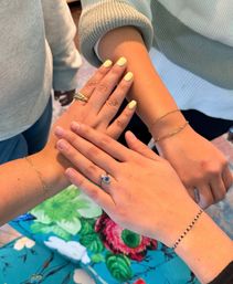 Three hands stacked over a colorful floral tablecloth showing gold bracelets, rings including a blue evil-eye charm, and one hand with pale yellow manicure.