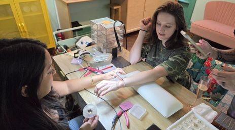 Woman having a delicate chain bracelet fastened by a technician at a beauty studio table with pliers, jewelry tray, nail tools and a champagne glass nearby.