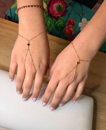 Delicate gold hand-chain bracelets with small red gemstones on both hands, pale pink manicure, resting on a wooden table with colorful floral fabric in the background.