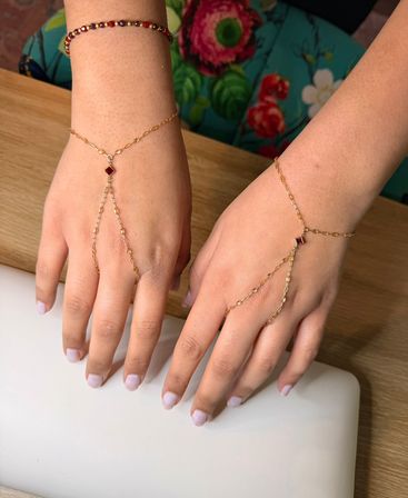 Delicate gold hand-chain bracelets with small red gemstones on both hands, pale pink manicure, resting on a wooden table with colorful floral fabric in the background.