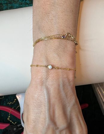Close-up of a forearm wearing two delicate gold bracelets — a dainty chain with a small opal bead and a layered chain with a tiny purple stone, jewelry close-up on a white display surface.