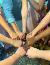 Circle of eight hands and wrists showing matching delicate bracelets, temporary "the bride" tattoos and colorful dresses — bridesmaid/bachelorette celebration over a patterned rug.