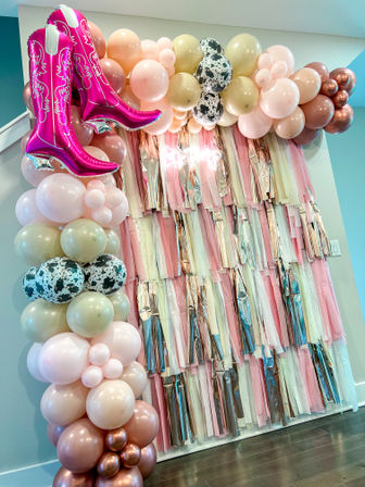 Indoor pastel party backdrop with blush, cream and rose-gold balloon arch, cow-print balloons and metallic pink cowboy-boot balloon over layered pink and rose-gold fringe photo booth wall.