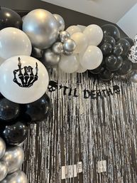 Indoor Halloween party backdrop with black, white and metallic silver balloon garland, a white balloon printed with a skeleton-hand, skull-themed "TIL DEATH" banner and shimmering silver fringe curtain.