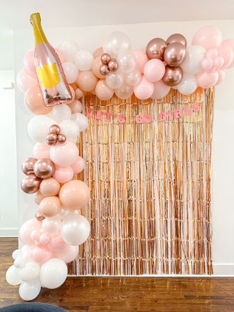 Indoor rose-gold fringe photo backdrop with blush pink, white, and metallic rose-gold balloon arch, oversized champagne bottle balloon, and banner staged on a hardwood floor for a girls' party photo-op