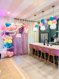 Indoor home birthday party in a modern kitchen with a pastel balloon arch and pink fringe backdrop, pink-striped ceiling, disco-globe lights and hanging balloons over a breakfast bar with wooden stools and festive place settings.