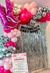 Vibrant pink-and-silver party photo backdrop with balloon arch, oversized pink cowboy-boot foil balloons, disco-ball and cow-print balloons, and a metallic fringe curtain.