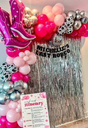 Vibrant pink-and-silver party photo backdrop with balloon arch, oversized pink cowboy-boot foil balloons, disco-ball and cow-print balloons, and a metallic fringe curtain.