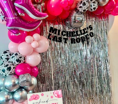 Vibrant pink-and-silver party photo backdrop with balloon arch, oversized pink cowboy-boot foil balloons, disco-ball and cow-print balloons, and a metallic fringe curtain.