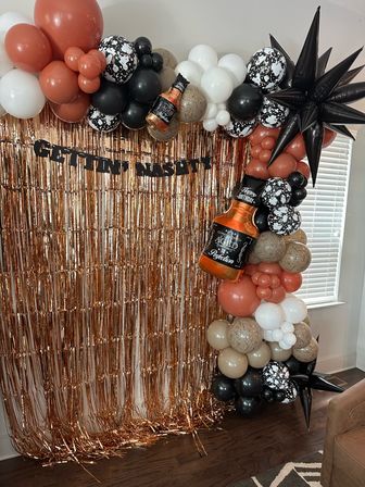 Indoor birthday photo backdrop with rose-gold fringe curtain and earthy balloon garland—terracotta, black, white and beige balloons with speckled accents, black starburst spikes and an inflatable whiskey-bottle foil balloon reading “Aged to Perfection”.