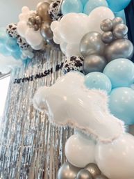 Indoor party photo backdrop featuring a pastel blue, white, and metallic silver balloon garland with speckled accents, a fuzzy cloud-shaped foil balloon, and a shimmering silver fringe curtain.