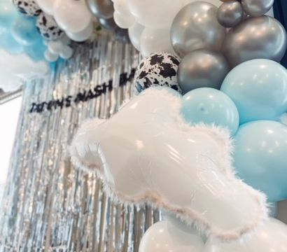 Indoor party photo backdrop featuring a pastel blue, white, and metallic silver balloon garland with speckled accents, a fuzzy cloud-shaped foil balloon, and a shimmering silver fringe curtain.