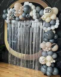 Stylish neutral balloon arch in gray, cream and white framing a silver fringe photo‑booth backdrop in a modern indoor event space, accented with bird silhouettes and a ‘love you’ scripted balloon.
