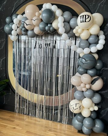 Stylish neutral balloon arch in gray, cream and white framing a silver fringe photo‑booth backdrop in a modern indoor event space, accented with bird silhouettes and a ‘love you’ scripted balloon.