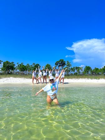 Cheerful group of friends in matching shirts splashing in clear shallow water at a white-sand beach with coastal pine trees and a bright blue sky