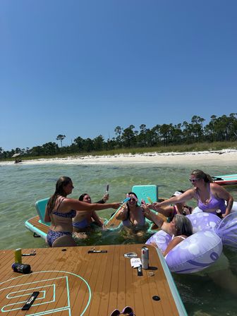 Group of women in swimsuits toasting with popsicles and drinks on paddleboards and a purple inflatable in clear shallow coastal water by a white sandy beach with pine trees under a bright blue sky