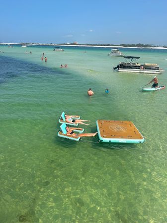 Sunny beach scene: people relaxing on inflatable floating loungers and a wooden-topped platform in crystal-clear turquoise shallow water, with pontoon boats, a paddleboarder and a distant sandy shoreline under a blue sky.