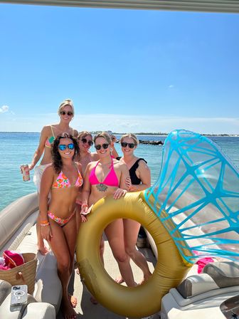 Five women in colorful bikinis and sunglasses posing on a pontoon boat with a large gold inflatable ring and blue canopy, sunny sky and clear turquoise coastal water