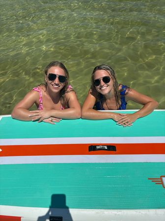 Two smiling people in sunglasses and swimsuits leaning on a colorful stand-up paddleboard in shallow green water on a sunny summer day
