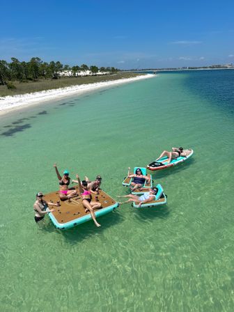 Group of sunbathers lounging on an inflatable floating dock and paddleboard in crystal-clear turquoise water off a white-sand beach under a sunny blue sky.
