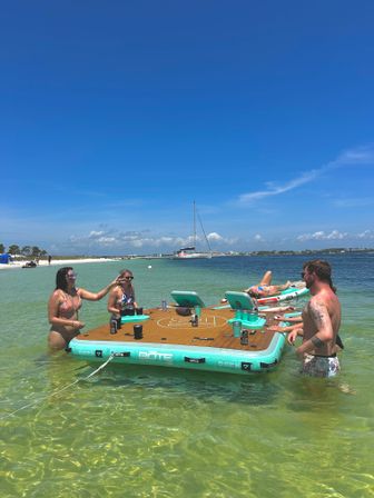 Group of friends in swimsuits enjoying a sunny beach day on a teal inflatable floating dock in shallow clear turquoise water, drinks on the platform and a sailboat anchored offshore under a bright blue sky.