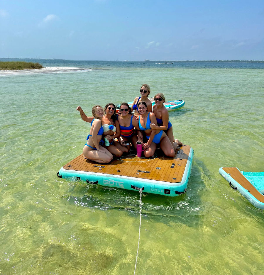 Six friends in colorful swimsuits sitting on a turquoise inflatable floating dock in shallow clear green water near a sandy beach with paddleboards nearby under a sunny blue sky