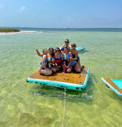 Six friends in colorful swimsuits sitting on a turquoise inflatable floating dock in shallow clear green water near a sandy beach with paddleboards nearby under a sunny blue sky