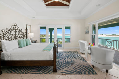 Bright oceanfront master bedroom with ornate iron headboard, white linens, turquoise accents, ceiling fan and balcony overlooking turquoise water and sandy beach.